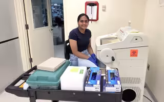 A young woman, seated, wearing blue jeans, a navy T-shirt, and blue latex glove, works at a machine in a biology lab. The machine is made of beige plastic and features a glass window on top and a circular crank on the side. A cart full of lab supplies is in front of her. 