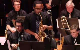 A group of musicians wear dark clothing and perform in a jazz ensemble with a saxophone player standing