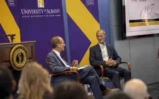 Two men sit in front of a purple backdrop with the words University at Albany on it while speaking before an audience of students and alumni.
