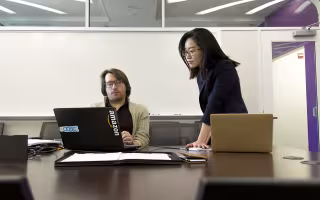 A professor sits at a desk before a computer as a student looks on