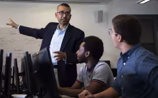 Professor Sanjay Goal gestures toward a whiteboard covered with diagrams while talking to two students seated at a table filled with computers.