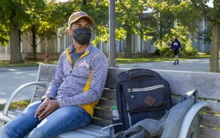 A student in a baseball cap and face mask sits on a park bench with his backpack