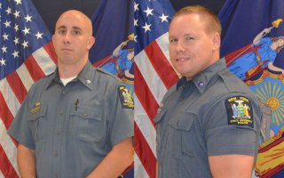 UUP Officers Daniel Callahan and Lucas Hoague stand in front of flags in their gray uniforms