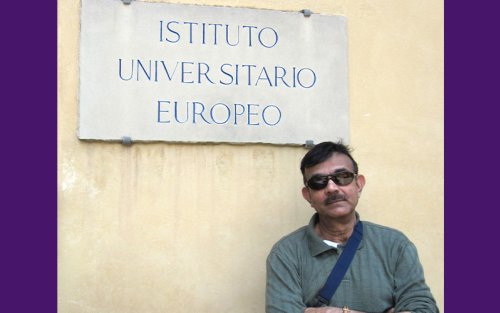 Kajal Lahiri stands with his arms crossed wearing a green long sleeved shirt and sunglasses. A blue bag strap falls across his chest and the bottom of the sign on the wall behind him reads "Instituto Universitario Europeo."