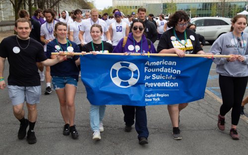 Photo depicts students leading UAlbany's 2023 "Out of the Darkness" walk. In front are six students holding a blue banner reading "American Foundation for Suicide Prevention, Capital Region New York". They are followed by a crowd of walkers. Brianna Lennon is among the banner holders, wearing a purple hoodie and dark aviator sunglasses. 