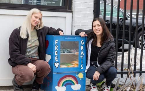 Two smiling young women crouch on a sidewalk next to a blue metal box containing period products. A rainbow and the words “Free” and “Gratis” are painted on the front of the box. A glass window displays period products inside. 