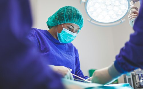 A surgeon wearing blue scrubs and a blue face mask works on a patient in a brightly lit room. 