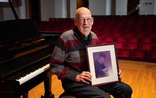 Professor George Hastings sits at a piano and holds a framed photo of his son Douglas.