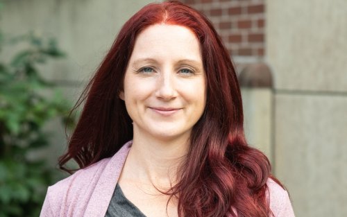 A smiling woman with red hair and a pink sweater stands outside in front of a stone building
