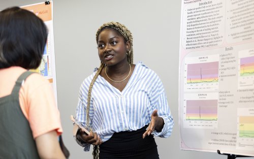An African -American young woman with a white and blue vertical-striped blouse, a gold necklace and a black skirt gestures with her hands while speaking in front of an academic poster indoors.