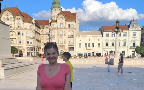 A woman in a pink blouse stands in front of a classical Roman large building in a city plaza with a blue sky and clouds in the background.