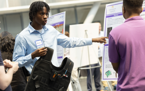 A man with short black braids in a light blue shirt holds up a black vest and gestures to a poster as he speaks to a man standing nearby in a purple shirt