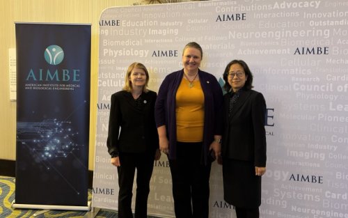 Three women smile for a photo, standing against a backdrop printed with the name “AIMBE” in purple, with a collection of science-related words in very faint print. To their left is a pull-up poster that says “AIMBE American Institute for Medical and Biological Engineering."