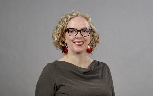 A profile photo of a woman with blonde hair, large red earrings and glasses wearing a dark brown blouse against a gray background.