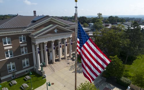 The American Flag is seen in the foreground on a flag pole in front of a colonial-style college building against a blue sky with green lawn and trees in the background.