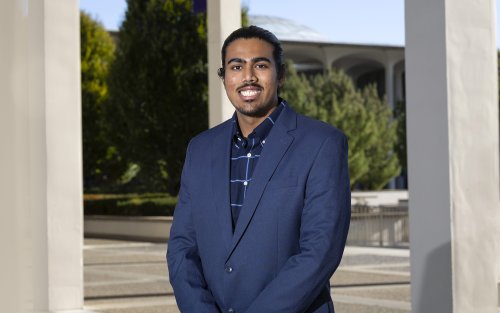 A young man stands outside amid a row of columns wearing a blue blazer and a blue shirt with trees and a dome in the background outside.