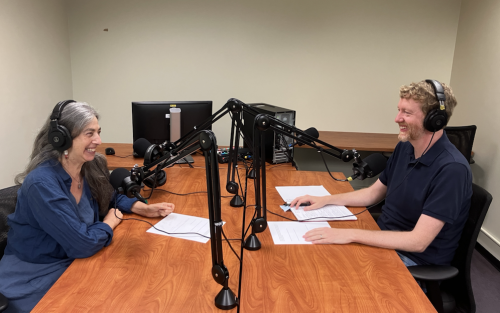 A man and a woman smiling in conversation as they are seated at a table in front of microphones for a podcast.