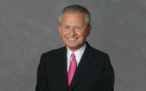 A man with silver hair in a black suit and pink tie smiles for a portrait against a gray backdrop.