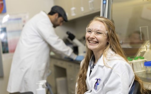 A woman with long wavy blond hair, wearing a white lab coat and clear protective goggles, smiles for a portrait in a lab. Lab benches with test tubes, and a man looking into a microscope, are in the background.
