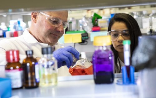 A photo of several glass bottles filled with colorful liquids with two scientists behind them in lab gear.