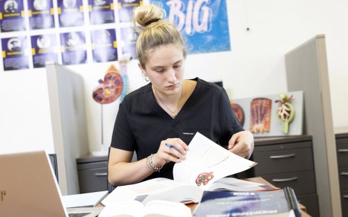 A young woman wearing black scrubs sits at a desk full of nursing textbooks. Anatomical models of a kidney are in the background. 