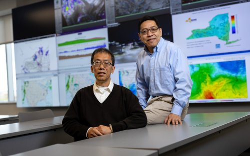 Professors Aiguo Dai and Liming Zhou sit at a table in front of weather maps at ETEC.