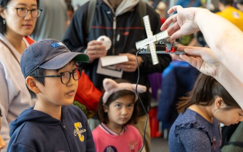A young boy in glasses and a babseball cap and a young girl in a headband watch as someone out of frame spins a windmill made from Legos.