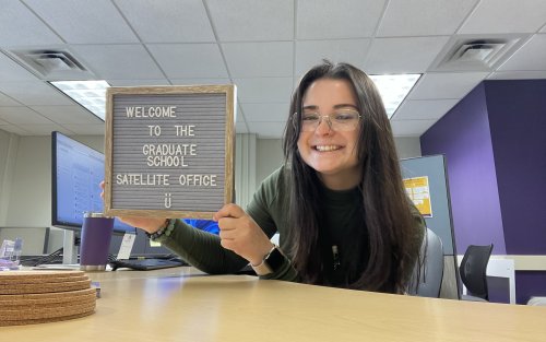 A woman holds a sign that says "Welcome to the Office," inviting guests into the workspace with a friendly gesture.