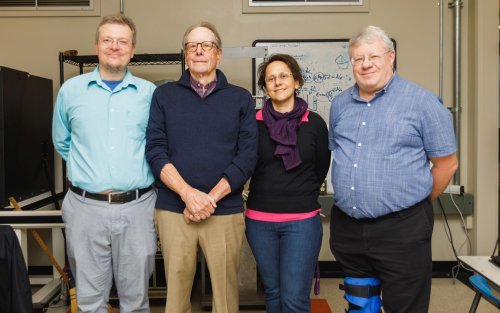 three men and a woman stand in a row facing the camera in front of a whiteboard scrawled with figures and shapes