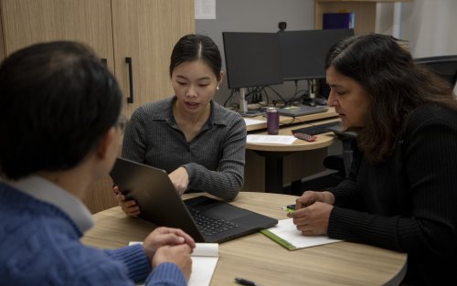 Three people sit around a laptop in an office as they go over something on the computer screen.