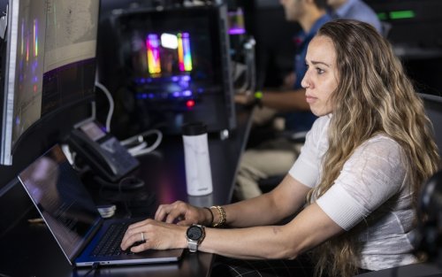 A woman in a white shirt sits at a laptop in a darkened computer lab with colorfully illuminated machines in the background. 