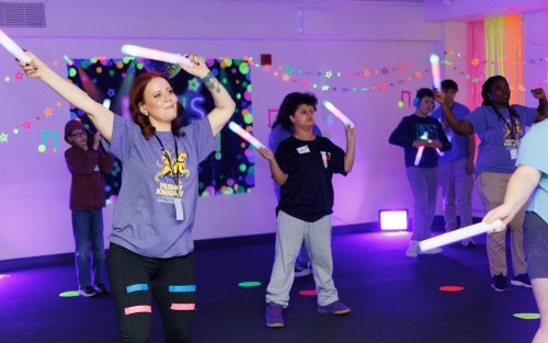 Children and adults wave glow sticks in a room that is lit purple and decorated with glow-in-the-dark stars and tape.