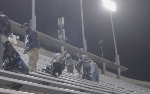 Six people carry trash bags, cleaning up stadium rafters at night under the stadium lights.