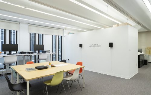 The installation space of Under-History Lessons. two speakers are mounted on a wall with the words "Under-History Lessons" on it in front of a table with chairs and office supplies.