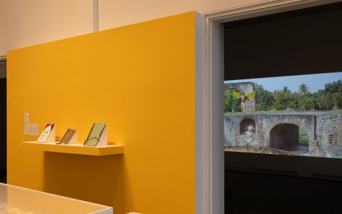 A yellow wall displaying some notebooks, behind the wall a projection plays.