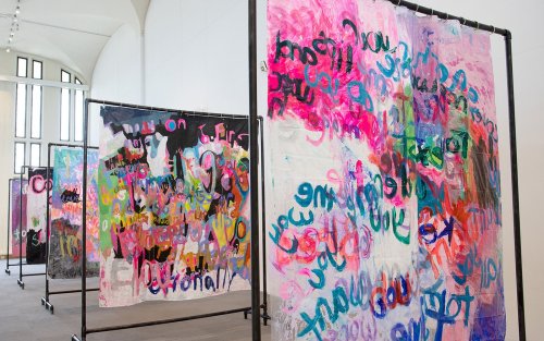A series of frames stand in the exhibition space, colorful shapes and words painted onto canvasses hanging from them.