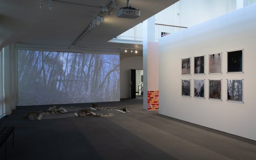 The exhibition space, 8 framed art pieces hang on a wall, with a projection playing onto a wall adjacent to it. Some rubble also sits on the floor.