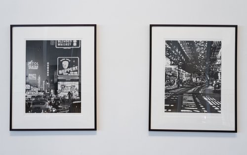 Two black and white photos hang on a wall, framed. They both depict urban scenes of a city.
