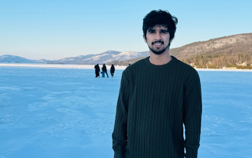 A man in a dark sweater stands on a frozen lake under a clear blue sky. Mountains line the horizon, with four distant people walking behind.