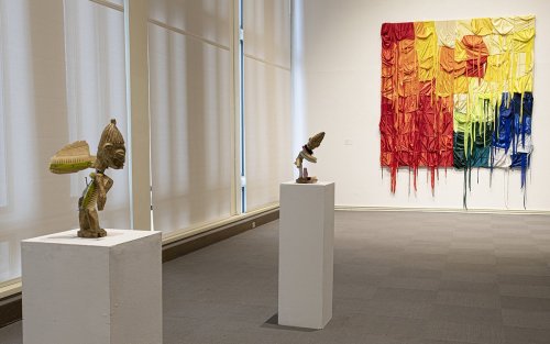 Two sculptures and a mounted artpice are displayed in the exhibition space. The scultpures are human-shaped, and the hanging piece is colorful and vibrant.