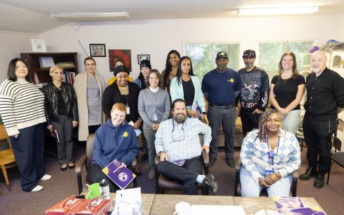 A group of sixteen people pose for a group portrait in a common area at Project Safe Point's Albany location. The room has multiple windows, artwork hung on the walls, and is lined in shelves containing books and bins of personal care supplies.