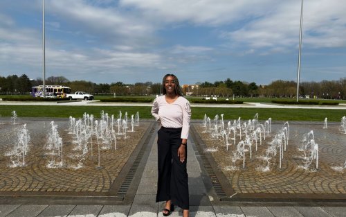 A person standing confidently near a fountain with water jets in a park, under a partly cloudy sky. Green grass and trees are visible in the background.