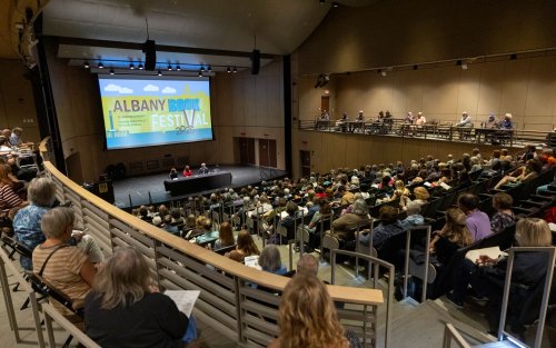 In a large auditorium people in stadium-style seating listen to a panel discussion on a stage at the bottom with a large visual display behind the panelists reads Albany Book Festival