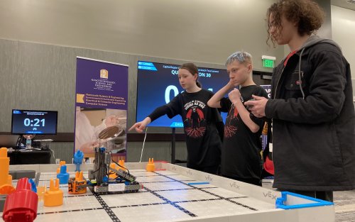 Girl points as two boys stare down at a robotic forklift on a game board.