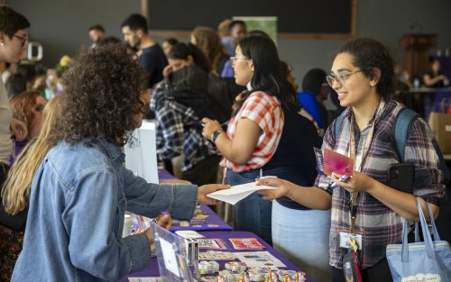 Two people interact at a busy indoor event table, exchanging papers, with informational materials and other attendees visible in the background.