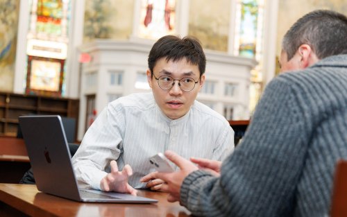 A man with dark hair and glasses sits at a table with a laptop in front of him and conversing with another man at the table holding a cellphone inside a picturesque library.