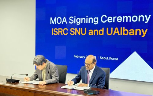 Two men sit a a brown desk signing documents in front of a blue screen that says "MOA Signing Ceremony"
