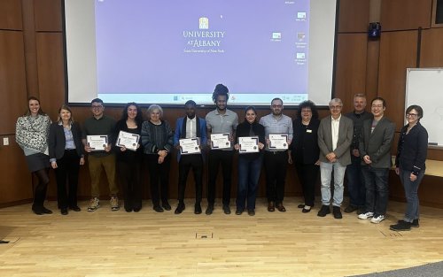 Fourteen people pose for a group portrait in front of a pull-down projection screen displaying the UAlbany logo. All are smiling and six are holding white paper certificates.