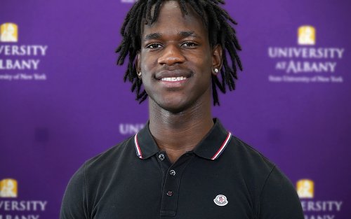 An African Americna young man wears a black collared shirt while standing against a purple backdrop with University at Albany written on it.