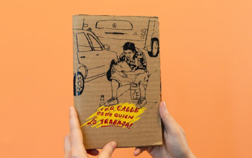 A person holds up a book. The title reads "¡La calle es de quien la trabaja!" and shows an illustration of a person sitting on a bucket with a car in the background.
